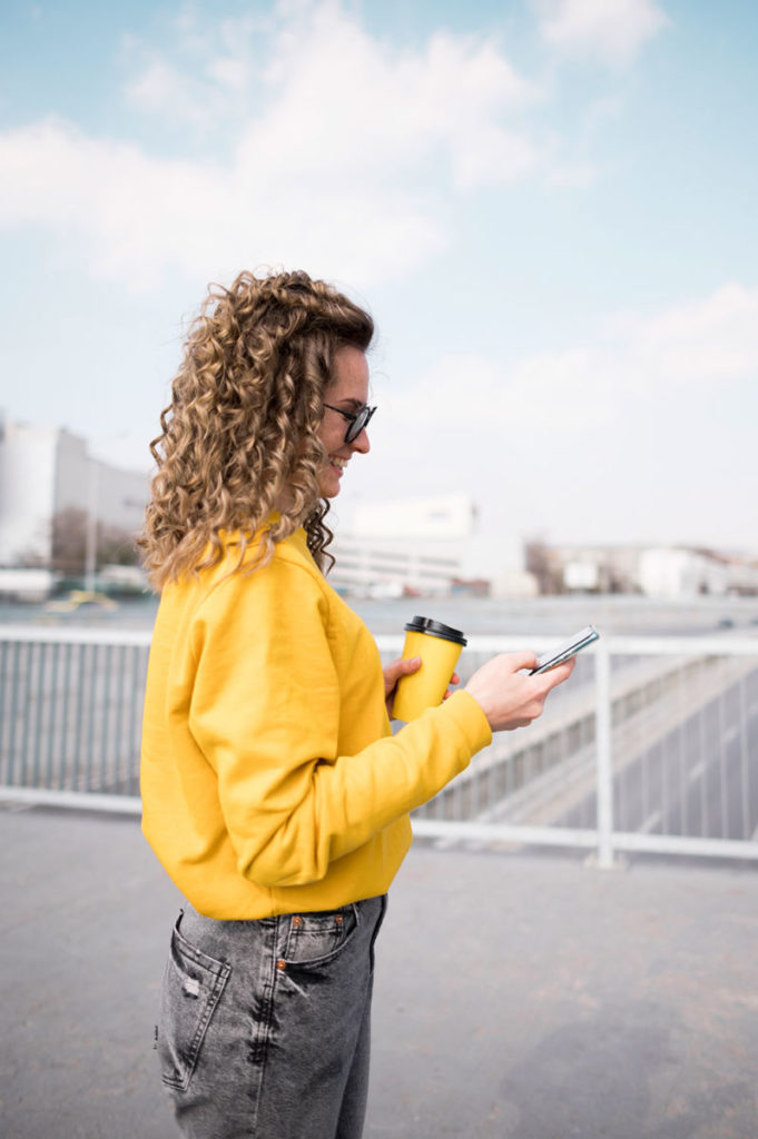 Person in yellow sweater with coffee and phone on urban rooftop, smiling under clear blue sky.